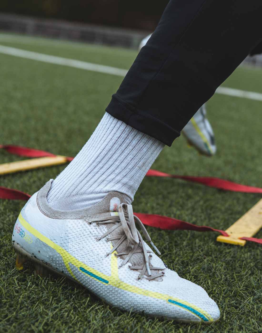 Nahaufnahme eines Fußballschuhs auf Kunstrasen. Vom Spieler ist eine weiße Socke und ein Teil der schwarzen Hose zu sehen. Im Hintergrund ist eine Koordinationsleiter und mehr Kunstrasen zu sehen.