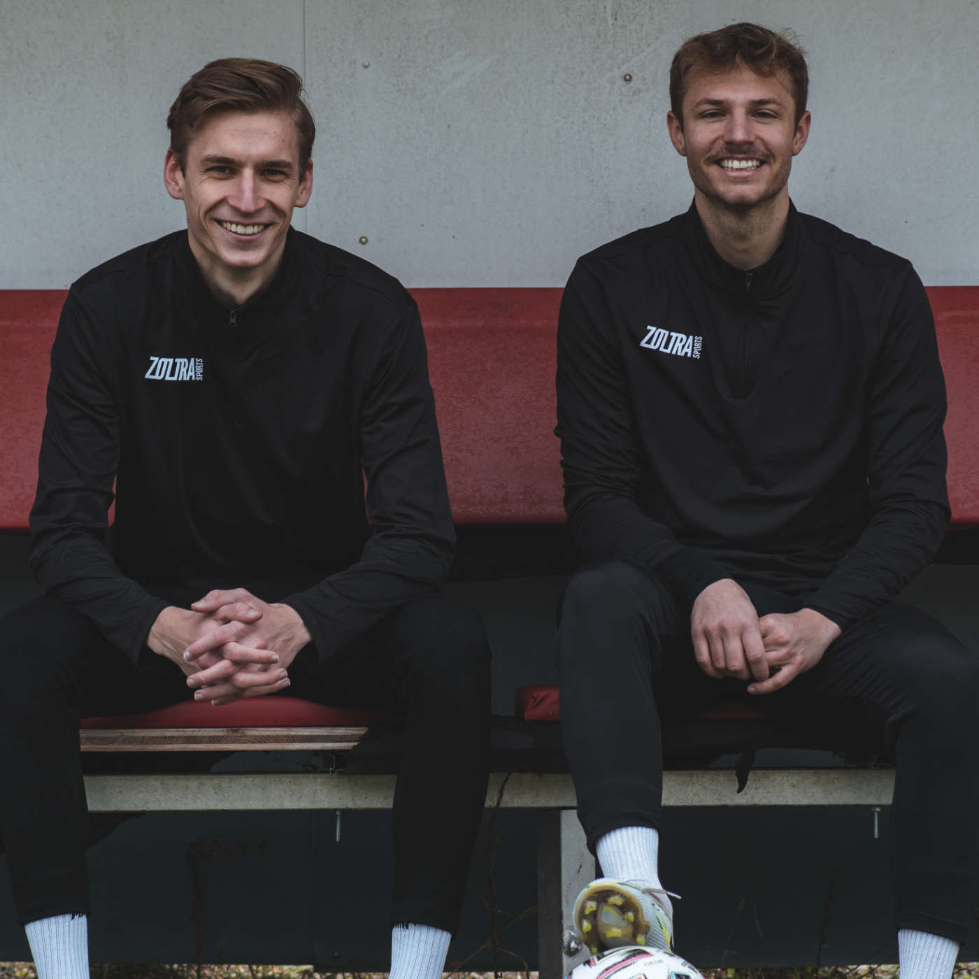 Die Gründer von Zoltra Sports, Mark (links) und Elias (rechts), sitzen auf einer roten Trainerbank. Beide tragen einen Zoltra Sports Pulli, Fußballschuhe und lachen in die Kamera.  Elias hat ein Bein auf einem Ball und Mark ist leicht nach vorne gelehnt.